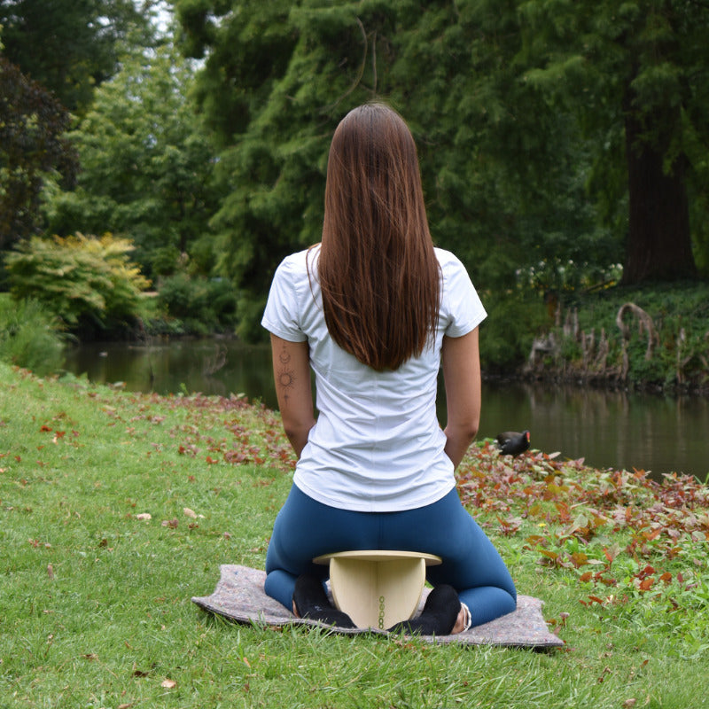 Une personne est assise en méditation au bord d’un étang, vêtue d’un t-shirt blanc et de leggings bleus, utilisant un banc de méditation noobeo.