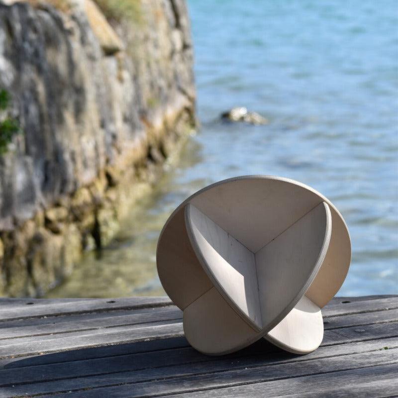 Le banc de méditation Noobeo est représenté renversé sur un quai en bois, avec ses deux supports en forme de demi-lune visibles sous l'assise ronde. À l'arrière-plan, on aperçoit une étendue d'eau calme et des rochers, sous un ciel clair.