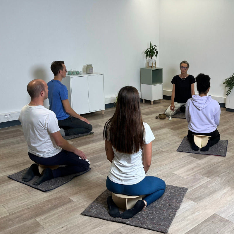 Cinq personnes sont assises sur des bancs de méditation Noobeo dans une pièce lumineuse et épurée. Ils sont disposés en cercle, dirigés vers une instructrice qui se tient au centre.